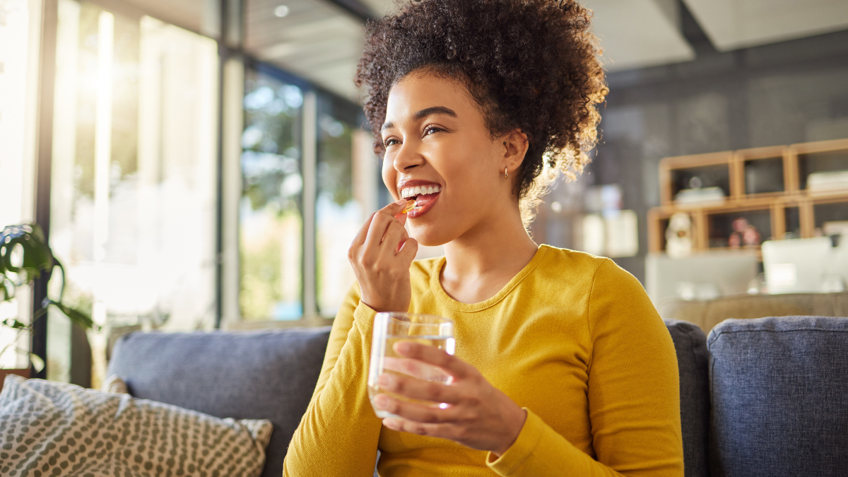 Young woman wearing a sweater and taking a vitamin with a glass of water while seating on the couch.